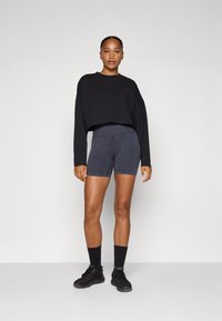 Felpa nera a maniche lunghe, abbinata a pantaloni da ciclismo gris scuro a vita alta. La modella indossa calzini neri alla caviglia e scarpe nere. Tessuto liscio, design aderente.