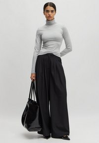 Grijze lange mouw turtleneck top gecombineerd met high-waisted zwarte wijde broek. Model houdt een glanzende zwarte tote bag vast.