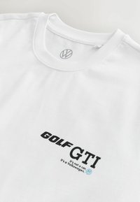 Hvid T-shirt lavet af blødt stof. Har sort "GOLF GTI" tekst og slogan nedenfor. Lille grå Volkswagen-logo på kraven.