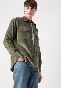 Katoenen korduroy overhemd in olijfgroen met twee borstzakken, knopen en een casual pasvorm. Gecombineerd met lichtblauwe denim jeans.