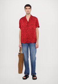 Rode t-shirt met korte mouwen en een luipaardprint, gecombineerd met lichtblauwe jeans en zwarte instapsandalen. Houdt een bruine papieren tas vast.