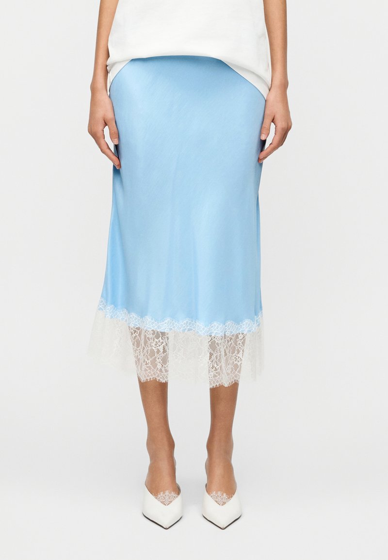 Φούστα μέσης μήκους από ανοιχτό μπ λε σατέν με λευκή δαντέλα στο τελείωμα. Διαθέτει λεία υφή και εφαρμοστή σιλουέτα. Φοριέται με λευκά παπούτσια μυτερά στην μύτη.