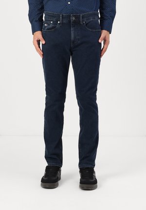 Homme portant un jean bleu foncé, des baskets noires et une chemise bleue à pois, debout devant un fond clair uni.