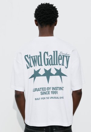 Férfi fehér pólóban, melyen a hátoldalon a "Stwd Gallery London" felirat és három csillag látható, fekete nadrággal párosítva.