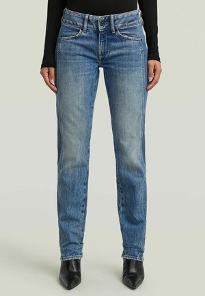 Personne portant un jean droit bleu, un haut noir à manches longues et des bottes noires à bout pointu, debout sur un fond uni.