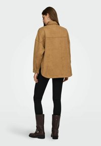 Suède tan jack met een relaxte pasvorm, achter schoudernaad en knoopmanchetten, gecombineerd met zwarte skinny jeans en bruine laarzen tot halverwege de kuit.