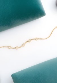 Guldarmband med hjärtformade länkar och klara rhinestones, mot en turkos och vit bakgrund. Delikat kedjedesign.