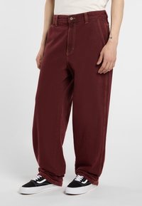 Pantalones de pierna ancha burdeos hechos de un tejido texturizado, con costuras amarillas, cierre de botón y bolsillos frontales. Llevados con zapatillas negras.