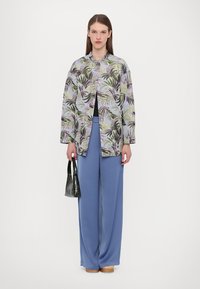 Camicia lunga e morbida con un motivo a foglie verdi e viola, indossata sopra una maglia nera e abbinata a pantaloni larghi blu chiaro. Borsa nera.