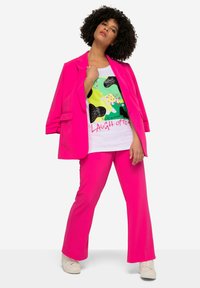 Ensemble composé d'un blazer rose vif et d'un pantalon assorti, associé à un t-shirt graphique blanc présentant des motifs abstraits verts et noirs. Des baskets blanches classiques viennent parfaire le look.