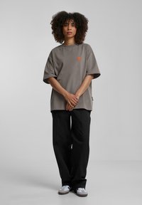 Camiseta gris de gran tamaño con logo naranja, de manga corta y un ajuste relajado, combinada con pantalones negros holgados y zapatillas casuales.