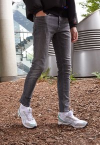 Harmaat farkut slim-fit-mallia yhdistettynä valkoisiin urheilukenkiin, joissa on vihreitä yksityiskohtia. Taustalla on luonnollinen maapinta ja moderni arkkitehtuuri.