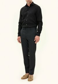 Zwart langemouwen shirt met een zak, gecombineerd met grijze op maat gemaakte broek en beige instappers met franjes, tegen een neutrale achtergrond.