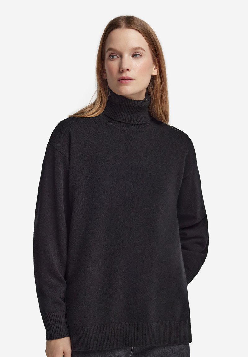 Frau mit heller Haut und langen, glatten Haaren, die einen schwarzen, übergroßen Rollkragenpullover trägt und leicht seitlich auf weißem Hintergrund schaut.