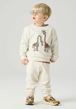 Bambino piccolo con capelli biondi e occhiali che indossa una felpa beige con stampa giraffa, pantaloni abbinati e sneaker gialle, che guarda di lato.