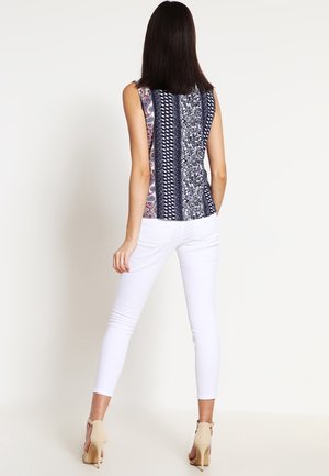 Frau mit glattem, dunklem Haar, die ein ärmelloses gemustertes Oberteil, weiße enganliegende Hose und beigefarbene High Heels trägt, steht mit dem Rücken zum Betrachter.