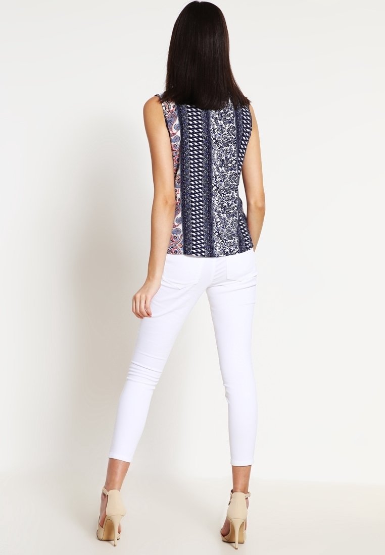 Nainen, jolla on suorat tummat hiukset, pukeutunut ilman hihoja olevaan kuvioituun toppiin, valkoisiin slim fit -housuihin ja beigeihin korkokenkiin, seisoo selkä katsojaan päin.