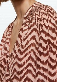 Primo piano di una persona che indossa una blusa rosa con motivi zigzag marroni e dettagli di tessuto arricciato sulla spalla.