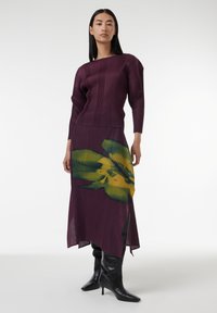 Top de punto de color ciruela con mangas largas, combinado con una falda fluida que presenta un estampado floral amarillo y verde sobre un fondo morado. Botas negras hasta la rodilla.