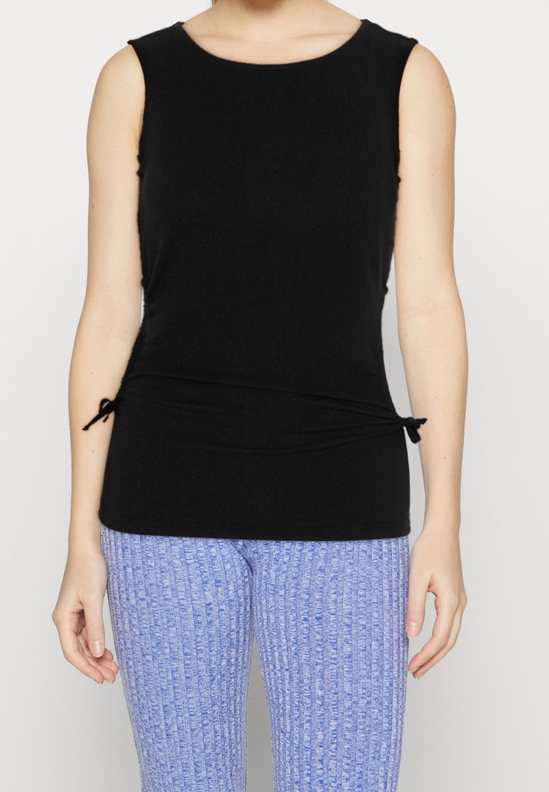 Femme portant un haut noir sans manches avec cordons latéraux et un pantalon lavande texturé, debout devant un fond clair uni.