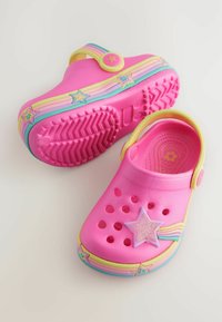 Ein Paar pinkfarbene Kinderclogs mit Regenbogen- und Sternverzierungen, belüfteten Löchern und verstellbarem gelbem Riemen auf hellem Hintergrund.