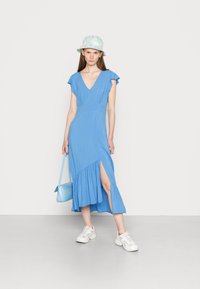 Vestido azul em decote em V com mangas curtas e esvoaçantes, fenda lateral e barra com babados. Combinado com uma mala azul clara e ténis brancos.