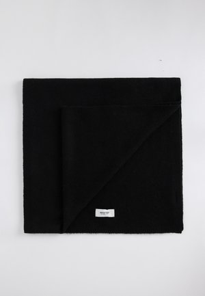 Couverture noire en tissu doux et texturé, pliée à un coin. Présente une petite étiquette blanche au bord inférieur.