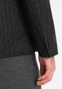 Blazer grigio scuro su misura con un tessuto testurizzato. Presenta tre bottoni neri sui polsini, abbinato a pantaloni grigi testurizzati.