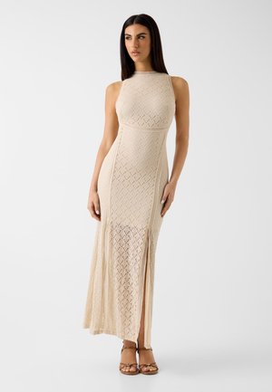 Donna con capelli lunghi e scuri che indossa un vestito maxi beige all'uncinetto senza maniche con spacco laterale e sandali marroni, in piedi su sfondo bianco.