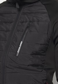 Black quilted jacket with vertical zip pocket on chest displaying white "JACK & JONES" logo.