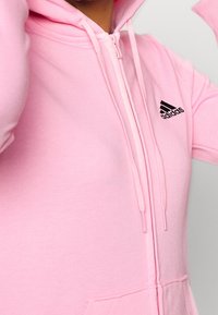 Sudadera con capucha de color rosa claro, con cremallera completa, logo de Adidas bordado en negro y cordones. Presenta una textura suave y un bolsillo canguro.
