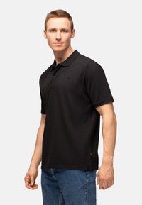 Polo nero realizzato in cotone, dotato di colletto, maniche corte e di un piccolo logo sul petto. Abbinato a jeans in denim blu.