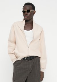 Ροζ απαλό τζάκετ με φερμουάρ και γιακά, με ελαστικά μανίκια, συνδυασμένο με λευκό τοπ και σκούρο γκρι παντελόνι με λεπτές ρίγες, αξεσουάρ με γυαλιά ηλίου.