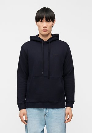 Fiatal felnőtt férfi egyenes fekete hajjal, egyszerű fekete kapucnis pulóvert és világoskék farmert viselve, fehér, egyszerű háttér előtt állva.