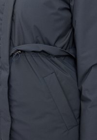 Veste matelassée bleu foncé avec ceinture, taille élastique et poche latérale, dotée d'une texture lisse et d'un léger éclat.