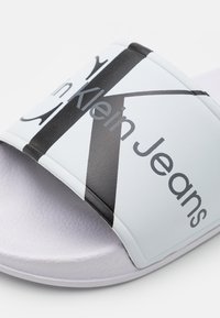 Sandal in pelle bianca con una larga fascia decorata da grafiche nere e testo. Il testo recita "in Klein Jeans." Soletta testurizzata per una migliore aderenza.