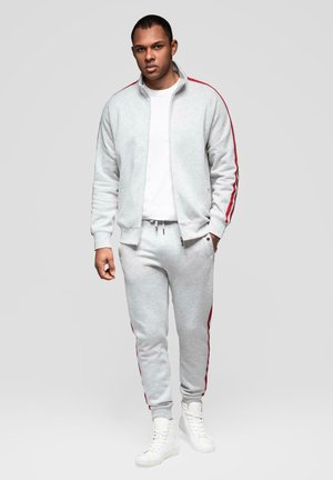 Homme debout, portant un survêtement gris clair avec des bandes rouges sur les manches et le pantalon, un T-shirt blanc et des baskets montantes blanches.