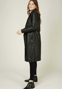 Manteau trench en cuir noir avec des revers et des fentes latérales, associé à des leggings noirs et des bottines à talons avec des accents de quincaillerie décoratifs.