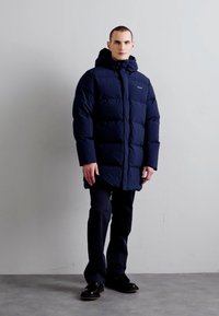 Marinblå puffjacka med huva, med en quiltad design, framsidan med dragkedja och en lätt glans. Kombineras med mörka byxor och svarta skor.