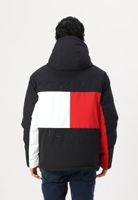 Veste isolée noire, rouge, blanche et bleue avec un grand logo, dotée d'un col haut et d'une capuche, affichant un design color block.