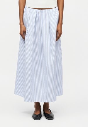 Donna che indossa una gonna lunga fino alla caviglia a righe verticali azzurro chiaro e bianco, abbinata a scarpe basse nere e una maglietta bianca.