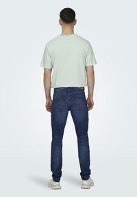 Lys grøn stribet kortærmet skjorte, mørkeblå jeans med baglommer og hvide sneakers. Set fra ryggen mod en lys baggrund.
