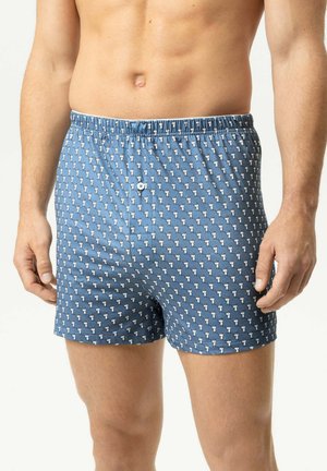 Mann trägt blaue gemusterte Boxershorts mit kleinen weißen Vogeldesigns, von der Taille bis zur Mitte des Oberschenkels vor einem einfarbigen hellen Hintergrund gezeigt.