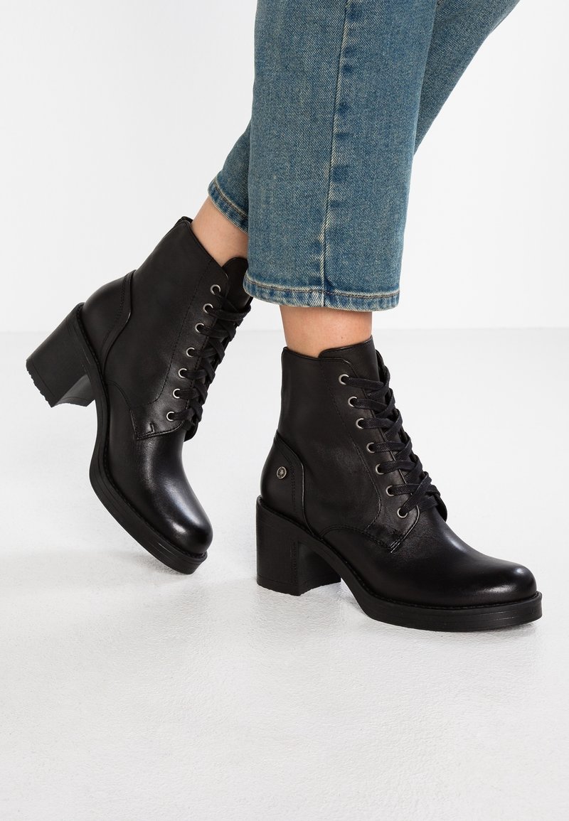 Bottes noires en cuir avec bout rond, fermeture à lacets, et talon épais. Le design présente une finition lisse et des accents discrets en métal.