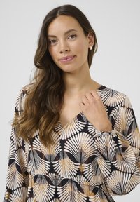 V-Ausschnitt-Bluse aus leichtem Stoff mit schwarzem Hintergrund und kontrastierenden weißen und goldenen Blattmustern. Elastische Bündchen und strukturierte Details.
