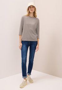 Gestreiftes Langarmoberteil in Grau und Weiß, kombiniert mit blauen Jeans. Das Model trägt helle Sneakers und eine helle Mütze. Einfaches, lässiges Outfit.