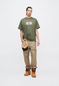 Olívazöld póló grafikus mintával, bézs színű cargo nadrág és barna munkacipő. A ruházathoz egy sötét peremű világosbarna sapka tartozik.