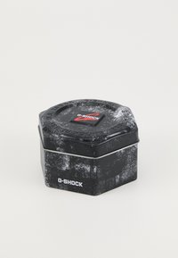 Heksagonal svart tinn med strukturert overflate, med en rød G-SHOCK-logo på toppen og synlig slitasjemønster; sikret med et sølvfarget metallbånd.
