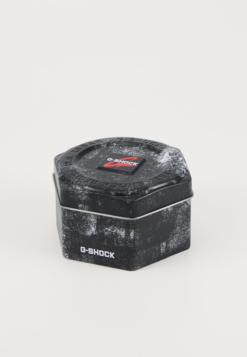 Scatola esagonale in latta nera con finitura testurizzata, caratterizzata da un logo G-SHOCK rosso in cima e un evidente schema di usura; fissata con una fascia metallica argentata.