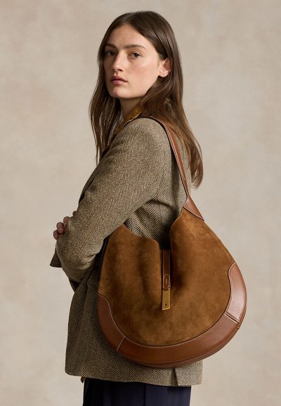 Ung kvinde med langt brunt hår iført en brun jakkesjakke med sildebensmønster, bærende en stor brun skuldertaske i ruskind og læder.